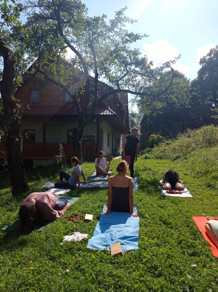 Grupa osób ćwiczy jogę na trawie, mają rozłożone maty. Wchodzą do pozcji supta virasana z blokiem.